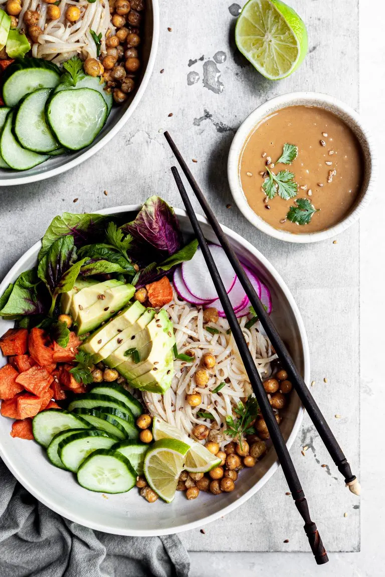 white bowl filled with rice noodles, cubes of sweet potatoes, chickpeas and cucumbers, with a small bowl of tahini dressing on the side