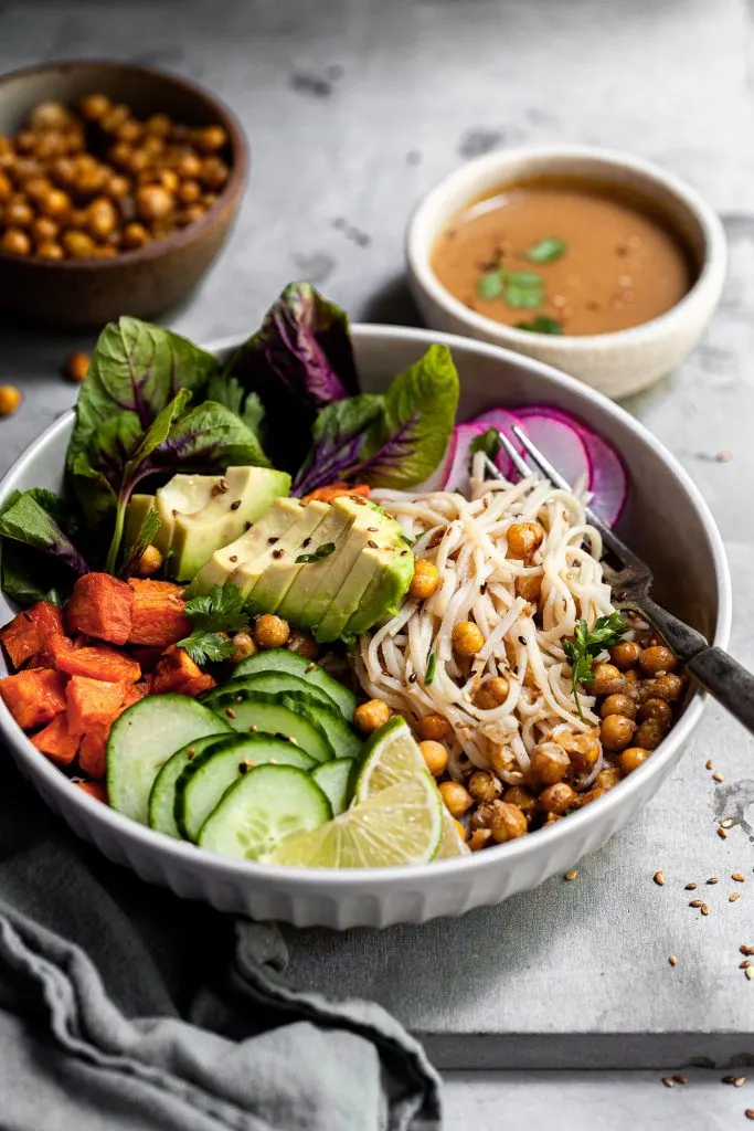 Side view into a bowl filled with rice noodles, roasted sweet potatoes, chickpeas, fresh veggies and sesame ginger sauce