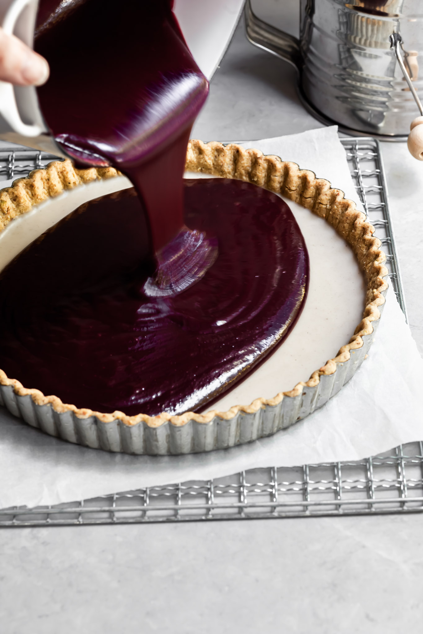 Purple berry curd is being poured into a tart crust already containing set white chocolate ganache