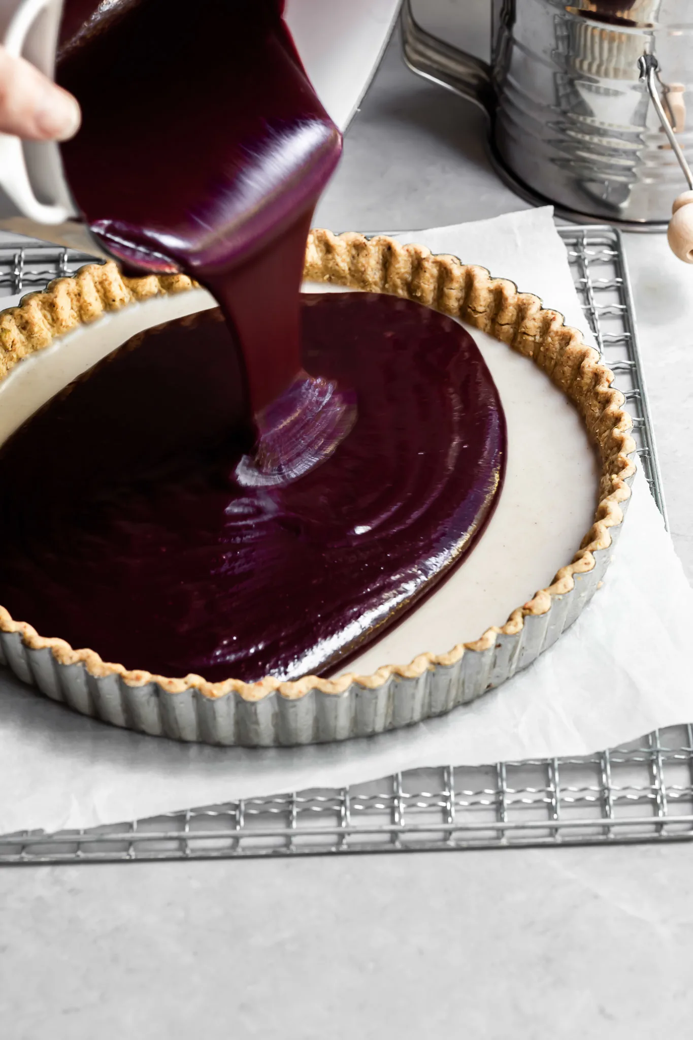 Purple berry curd is being poured into a tart crust already containing set white chocolate ganache
