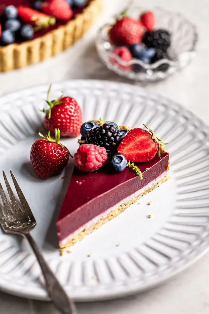 Slice of a layered tart - bottom is shortbread lined with white chocolate and then a layer of purple berry curd. Garnished with berries, served on a white plate. 