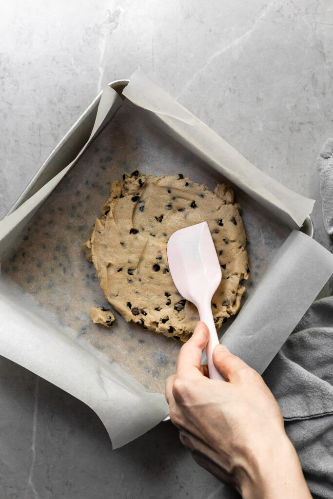 2 layers of chocolate chip cookie dough being pressed into a square aluminium tin. The layers are separated by a sheet of parchment paper. 