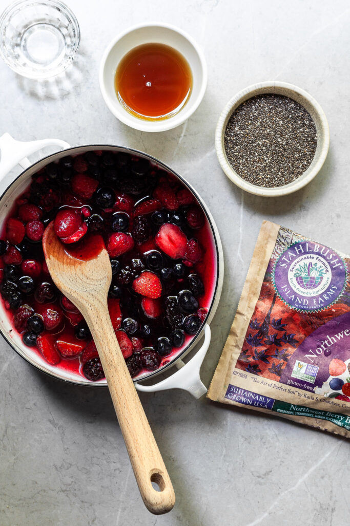 a white pot filled with a berry blend and water, with a small bowl of chia seeds, another containing maple syrup and another filled with vanilla extract