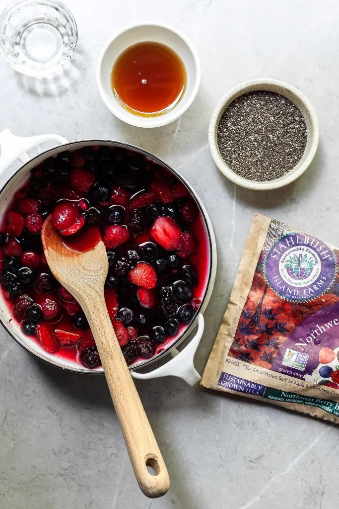 a white pot filled with a berry blend and water, with a small bowl of chia seeds, another containing maple syrup and another filled with vanilla extract