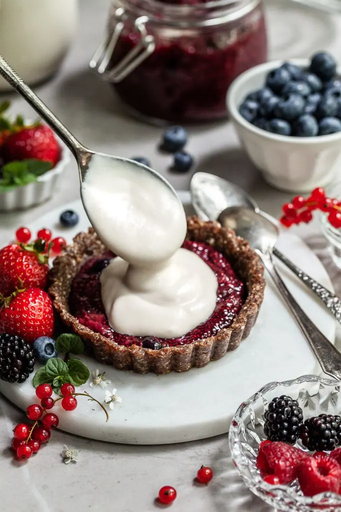 A tart shell with a layer of berry chia jam is being topped with vegan yogurt