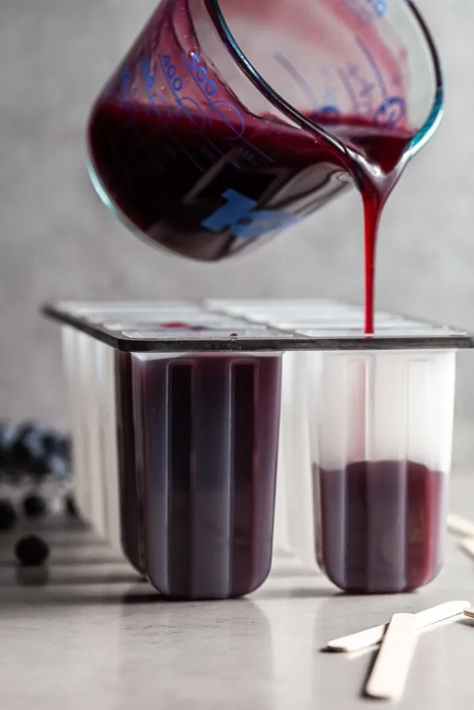 Rich purple syrup being poured into a popsicle mold from a glass measuring cup