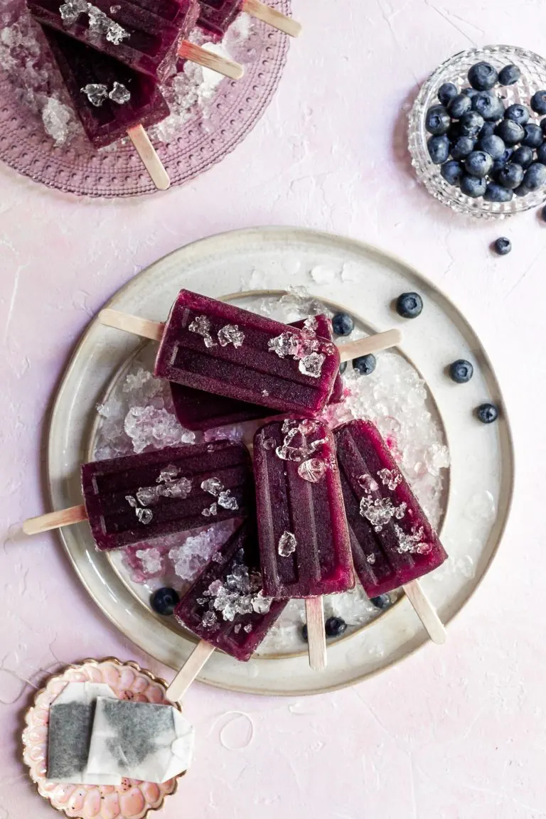 a cream colored plate covered in crushed ice with purple popsicles and blueberries resting on top