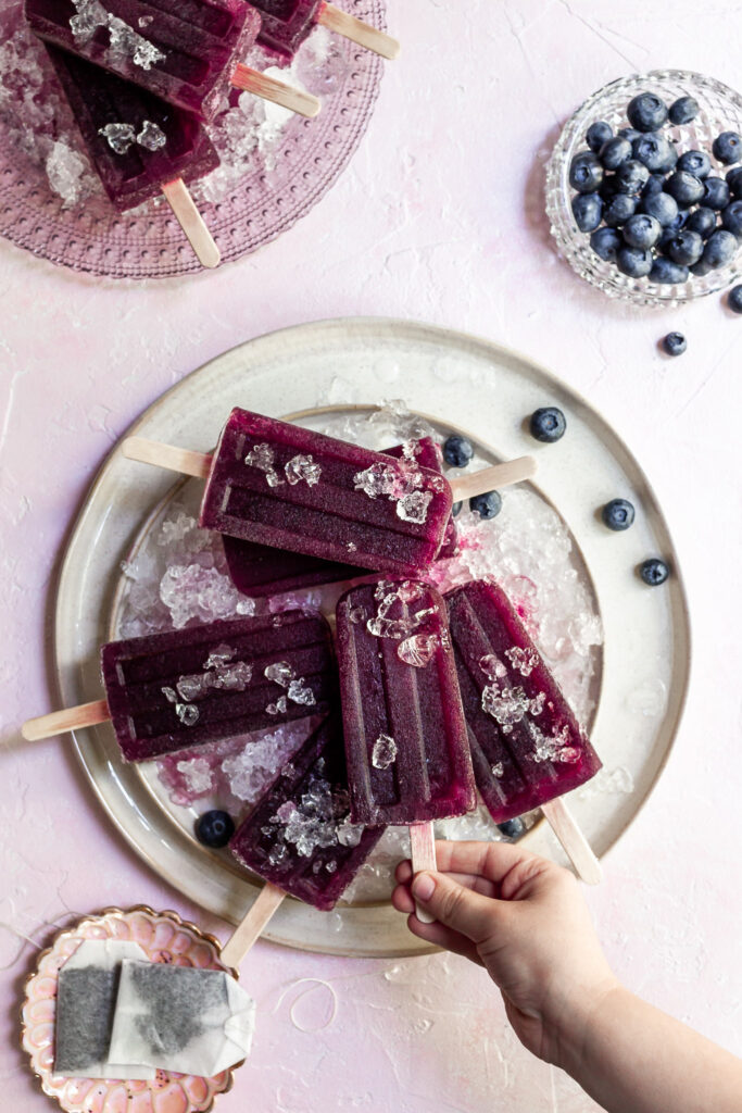a small child's hand grasps the stick of a purple popsicle from a cream serving plate covered in crushed ice