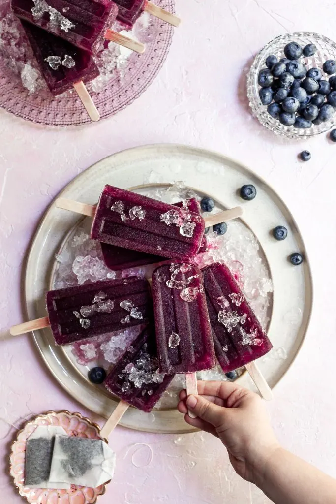 a small child's hand grasps the stick of a purple popsicle from a cream serving plate covered in crushed ice