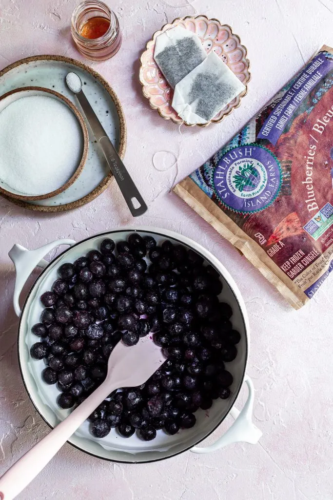 Preparation for making blueberry early grey popsicles. A white pot filled with frozen blueberries, beside a bowl of sugar and 2 earl grey tea bags.