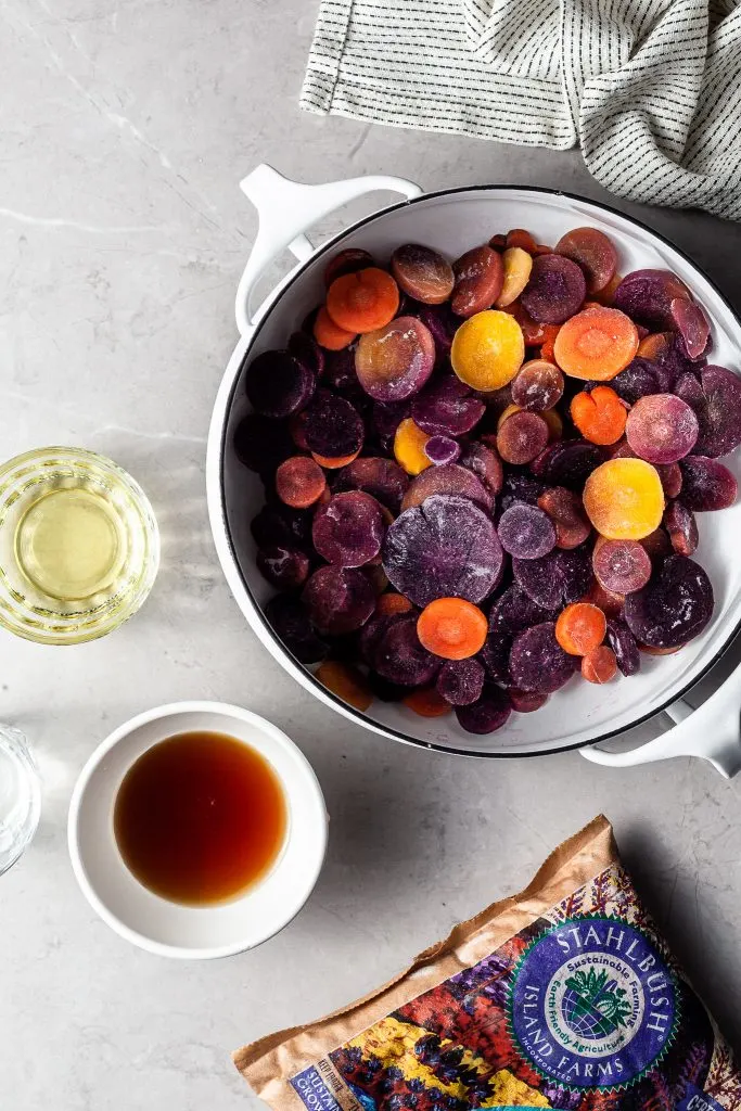 White pot filled with frozen sliced tri-color carrots, beside a small bowl of maple syrup 