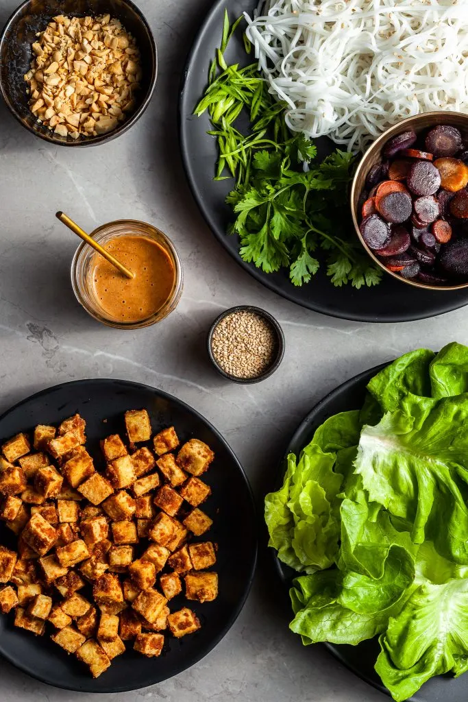 the ingredients prepped in bowls for assembling the lettuce wraps - cooked rice noodles, herbs, glazed carrots, cubes of tofu, peanut sauce and lettuce leaves.