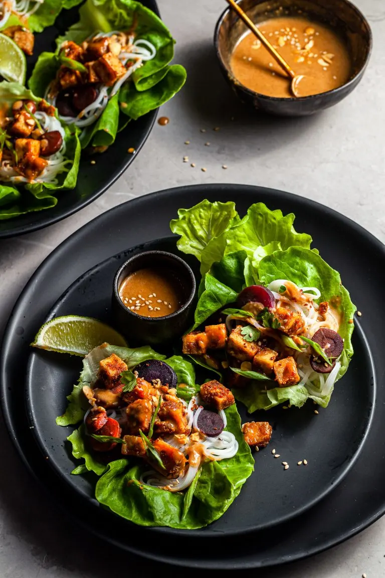 lettuce leaves on a black plate filled with tofu, rice noodles, carrots and drizzled with peanut sauce