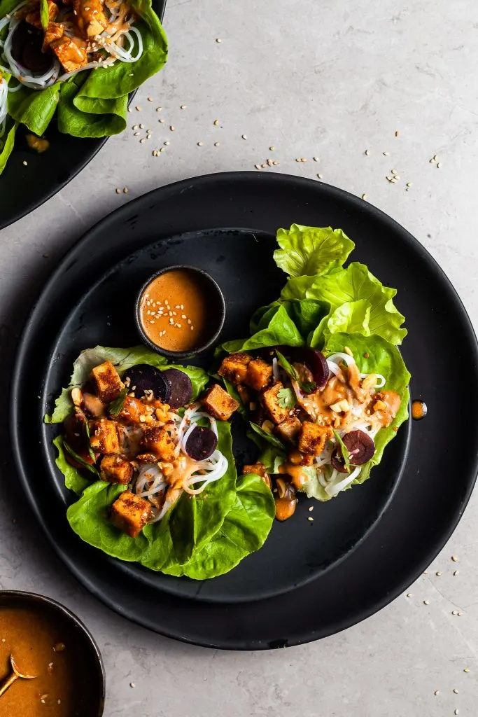 looking down on a black plate filled with 2 lettuce leaves filled with tofu, rice noodles, carrots and drizzled in peanut sauce