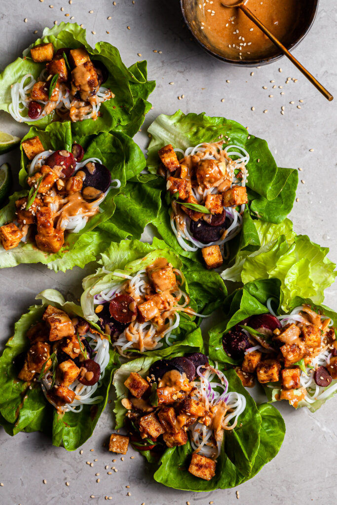 lettuce leaves on a marble counter top filled with cubed tofu, rice noodles, and carrots drizzled in golden brown sauce