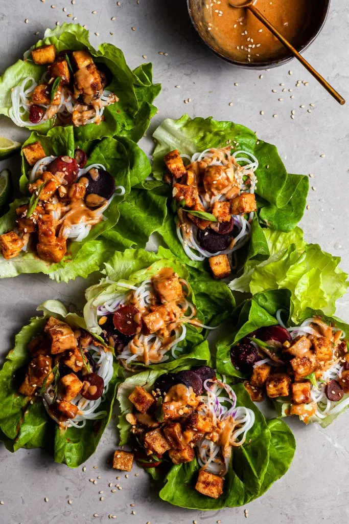 lettuce leaves on a marble counter top filled with cubed tofu, rice noodles, and carrots drizzled in golden brown sauce