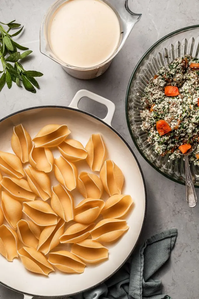 Components for the butternut squash stuffed pasta on a marble counter: jumbo pasta shells, a vegan ricotta/sausage/roasted butternut squash mixture and a butternut squash cream sauce
