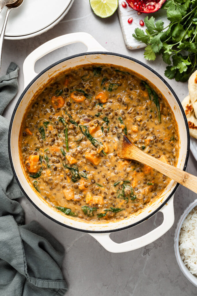 A white enamel cast iron pot filled with sweet potato lentil curry. Beside the pot are serving bowls, cooked rice and garnishes like lime wedges and cilantro