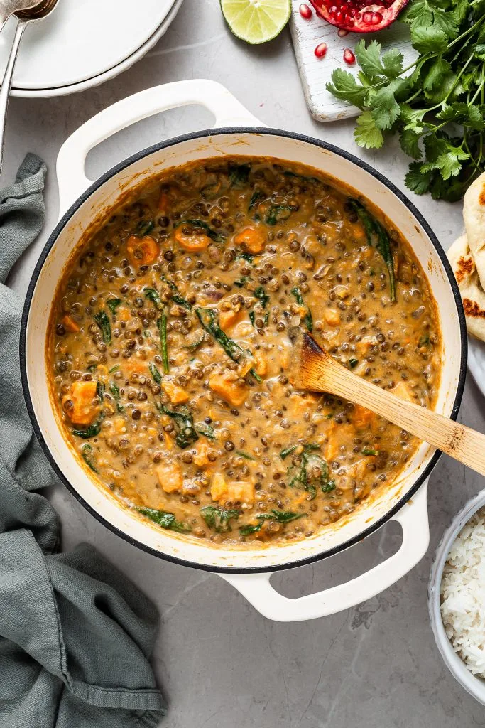 A white enamel cast iron pot filled with sweet potato lentil curry. Beside the pot are serving bowls, cooked rice and garnishes like lime wedges and cilantro