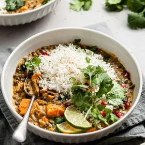 White bowl filled with sweet potato lentil curry garnished with cilantro, slices of lime, and pomegranate arils