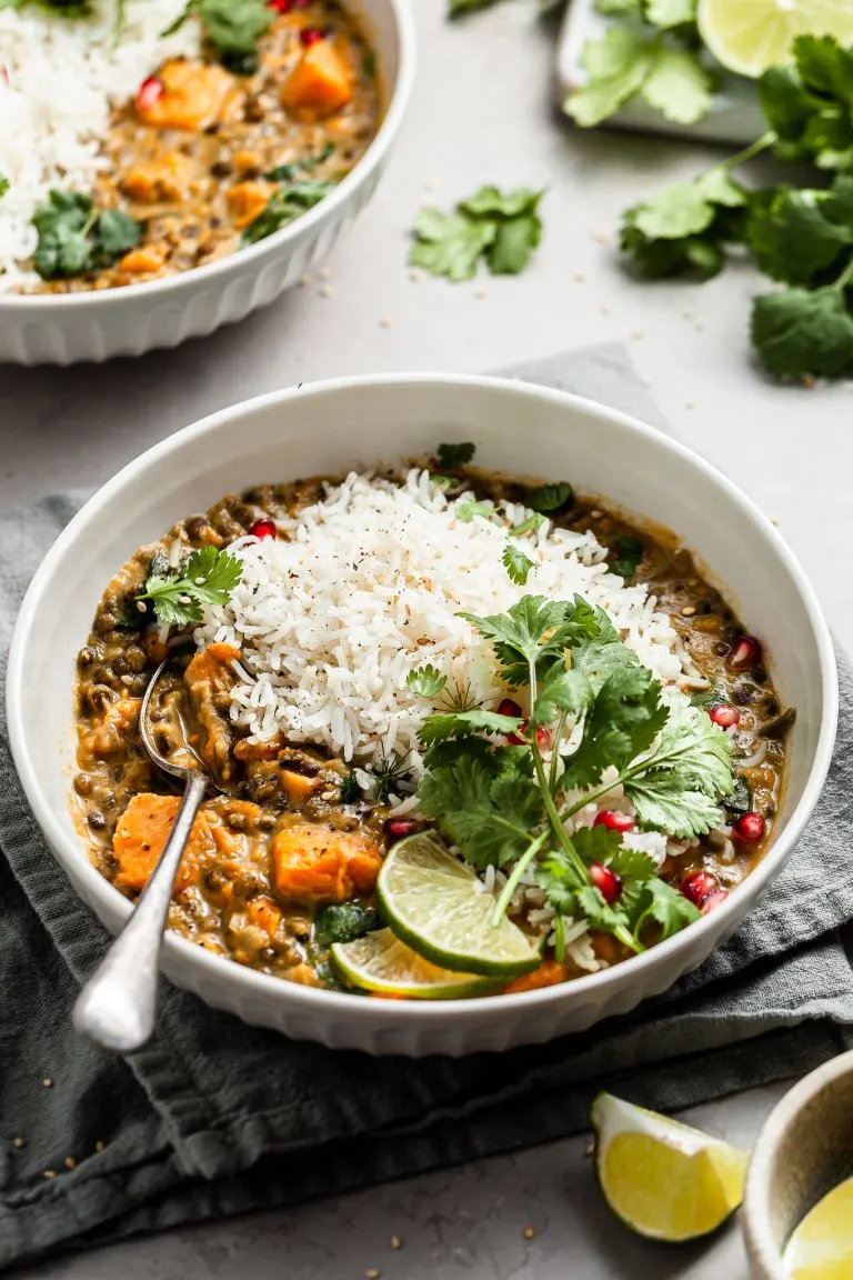 White bowl filled with sweet potato lentil curry garnished with cilantro, slices of lime, and pomegranate arils