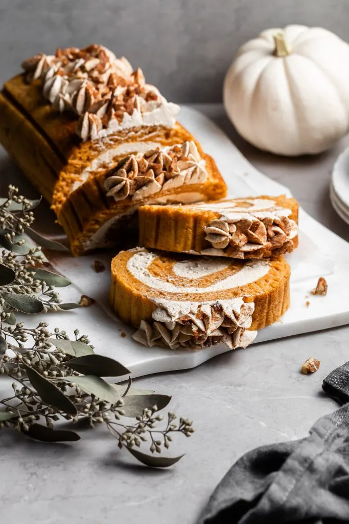 A pumpkin dessert roll resting on a white platter with three slices to be served and white plates at the side