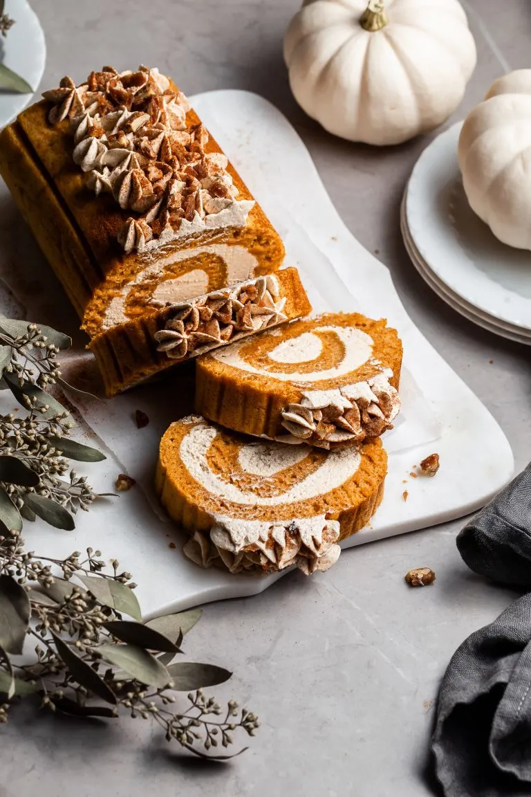pumpkin roll on a white plater beside 2 small white pumpkins