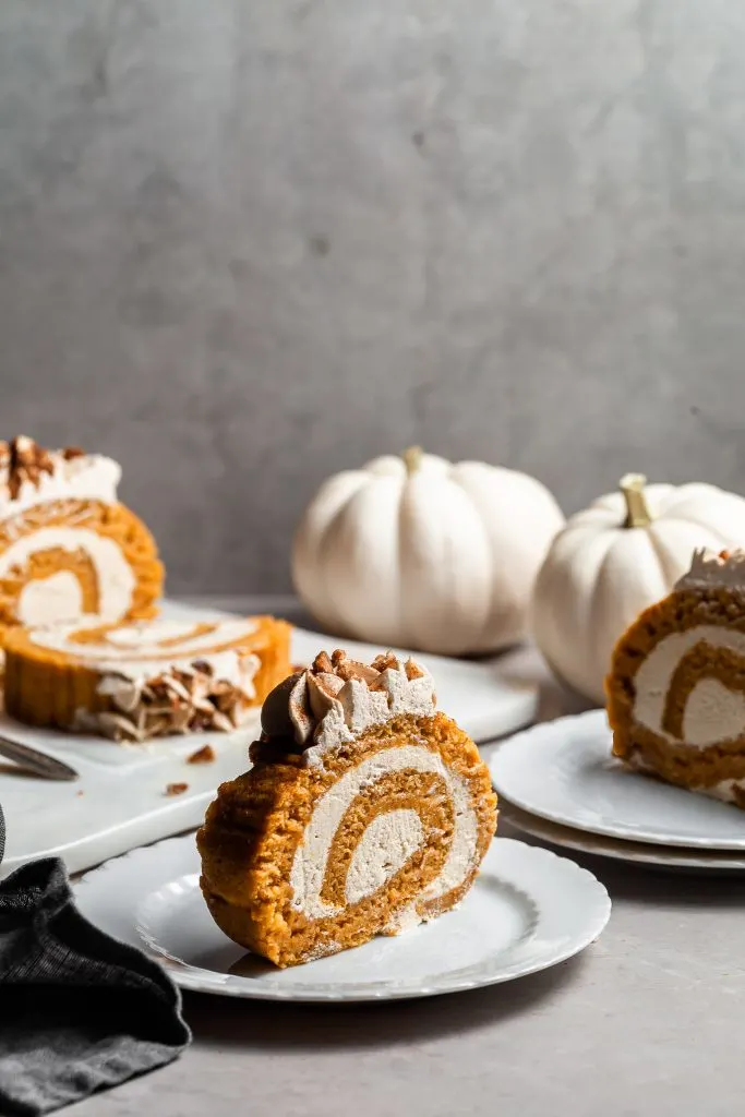 Slices of pumpkin roll on white plates in the foreground and the sliced pumpkin roll in the background