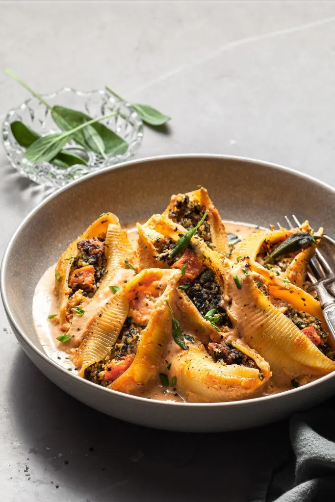 Stuffed pasta shells in a grey bowl with light pouring into the scene from behind a small bowl of fresh sage leaves
