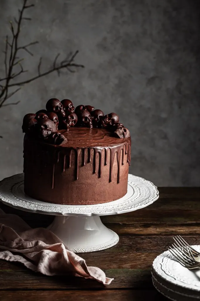 Chocolate layer cake on a white cake stand. A stack of white plates is in the foreground on top of which lies 3 silver forks. 
