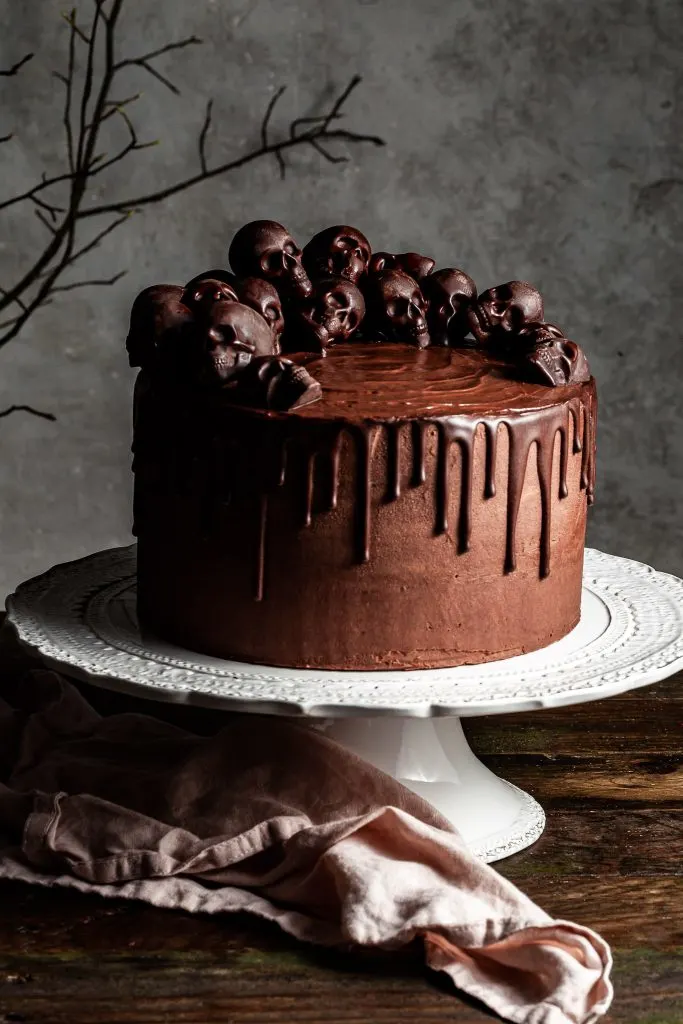Chocolate layer cake with ganache drips and a half crown of small chocolate skulls on the top. The cake is on a white cake stand with a pink linen napkin beneath it. 