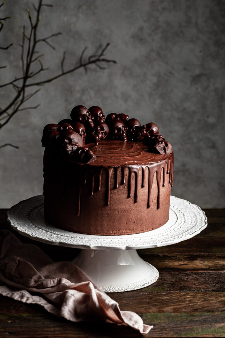 Chocolate layer drip cake decorated with small chocolate skulls, sitting on a white cake stand
