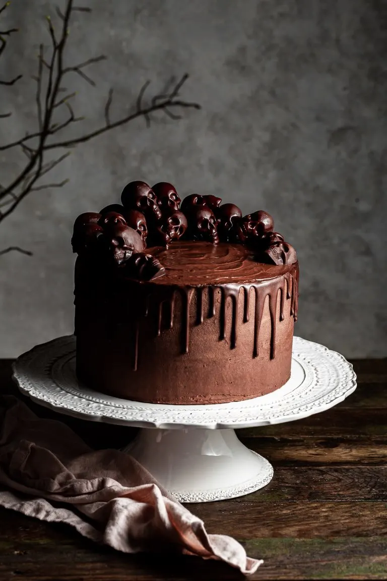 Chocolate layer drip cake decorated with small chocolate skulls, sitting on a white cake stand