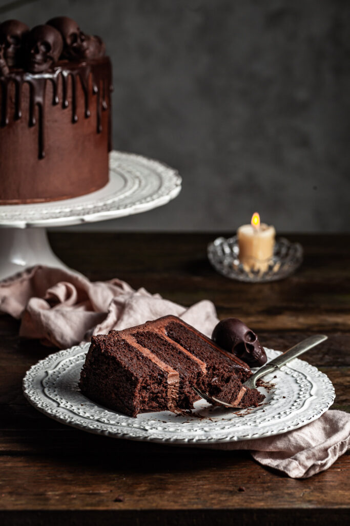 View of a slice of a triple layer chocolate cake with several bites missing and a fork resting on the plate it's lying on. A chocolate skull sits on the plate, while a candle glows in the background. 