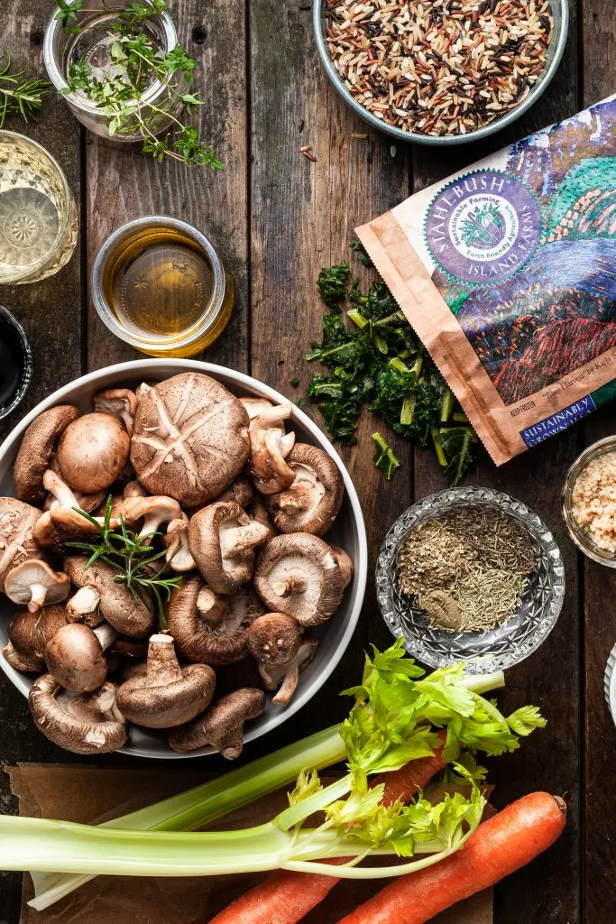 Flatlay of the ingredients to make the mushroom rice soup, including a bowl of mixed mushrooms, carrots, celery, herbs, minced garlic and a bag of frozen chopped kale