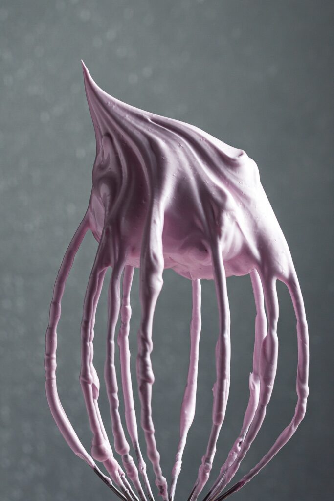 A balloon whisk from a stand mixer held upside down covered in pink meringue