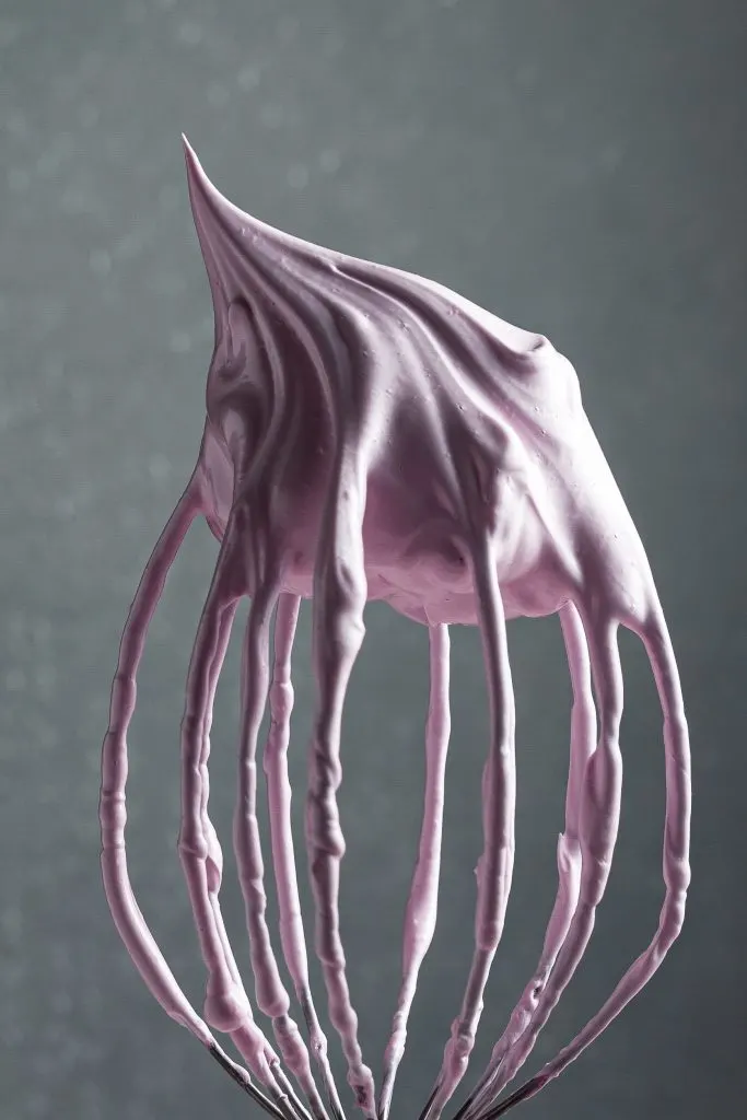 A balloon whisk from a stand mixer held upside down covered in pink meringue