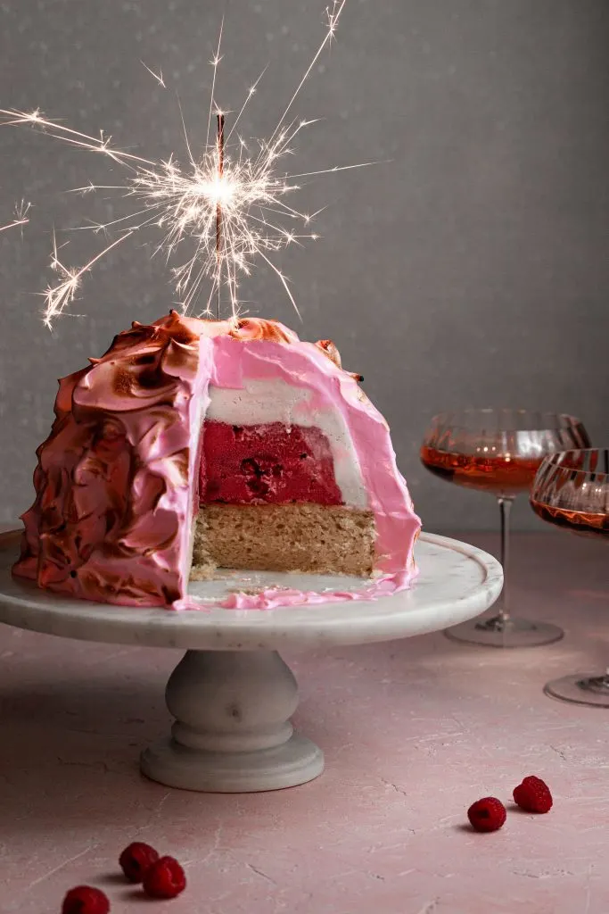 Pink baked Alaska with a lit sparkler on top beside 2 glasses of wine