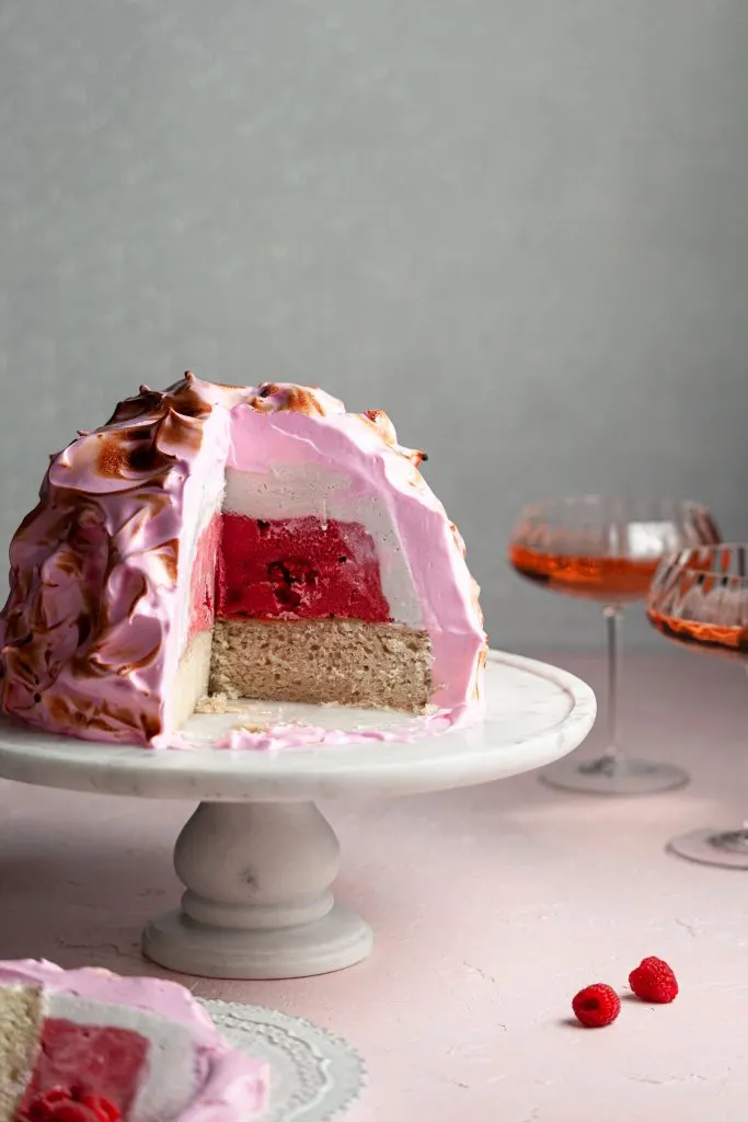 A slice of baked alaska in the foreground, in front of a baked alaska covering in meringue sitting on a marble stand