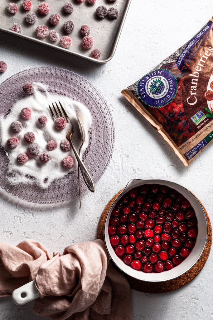 A view of sugared cranberries being made. A pot of cranberries in syrup at the bottom of the scene, with a glass plate filled with white sugar and a few cranberries already rolled in the sugar, a bag of frozen cranberries and a small baking sheet with sugared cranberries drying. 
