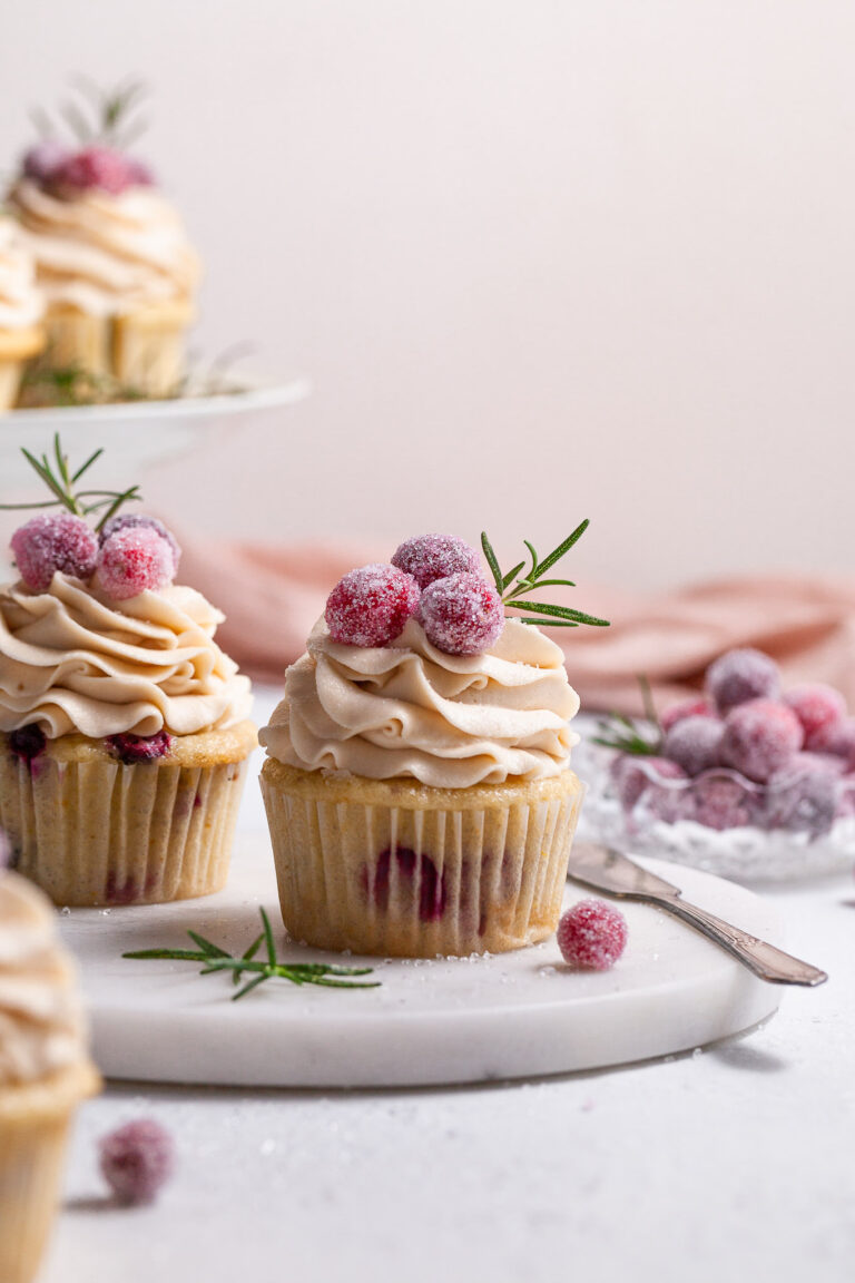A cranberry cupcake topped with piped frosting is garnished with candied cranberries , sitting on a marble plate