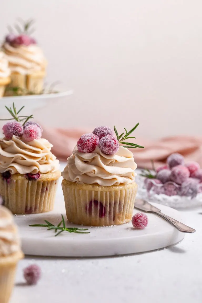A cranberry cupcake topped with piped frosting is garnished with candied cranberries , sitting on a marble plate