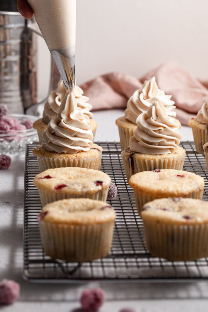 Cupcake bases on a silver cooling rack where a fifth one has just been frosted with the piping tip just lifting. Other bases have yet to be frosted. Sugared cranberries lie on the counter. 