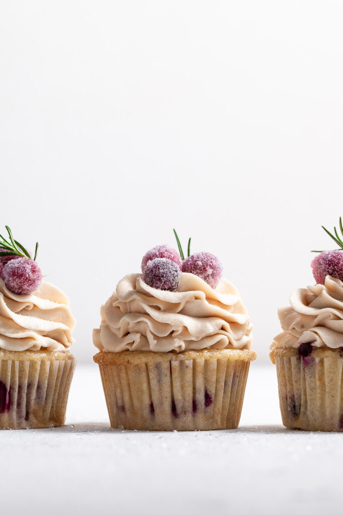 Eye level view of 3 frosted cupcakes, the bases appear to have red berries in the cake. The frosting is pipe high and topped with candied cranberries and a sprig of rosemary each.
