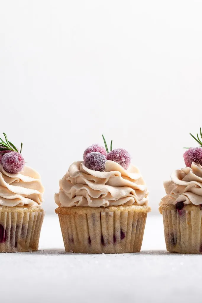 Eye level view of 3 frosted cupcakes, the bases appear to have red berries in the cake. The frosting is pipe high and topped with candied cranberries and a sprig of rosemary each.