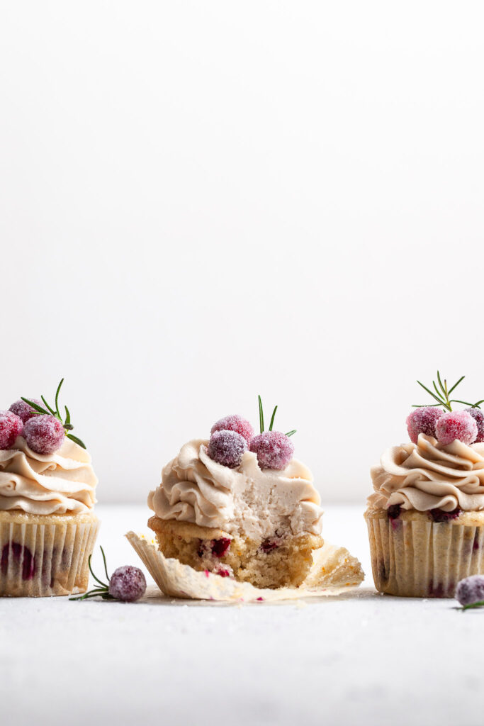 Three frosted cupcakes in a row at eye level, garnished with candied cranberries. The center cupcake has a bite missing. 