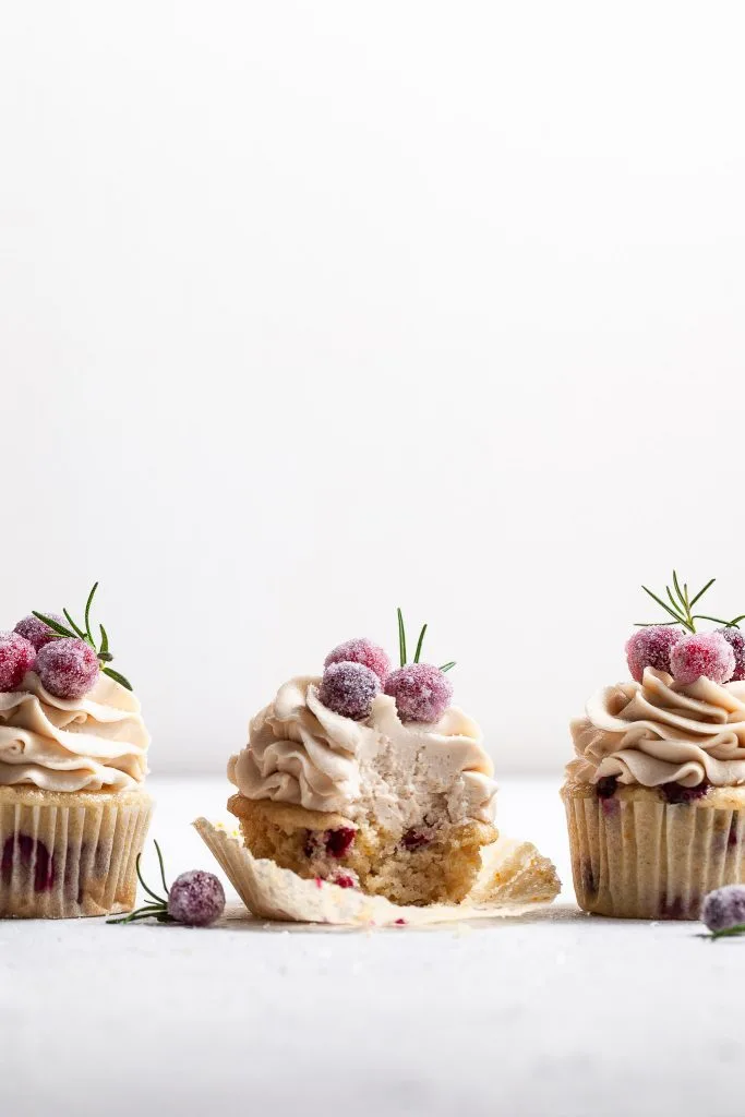 Three frosted cupcakes in a row at eye level, garnished with candied cranberries. The center cupcake has a bite missing. 