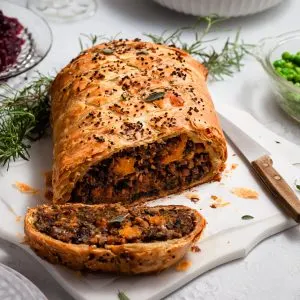 A vegan Wellington on a white marble platter sitting on a tabelscape with a bowl of whipped potatoes, green peas, cranberry sauce and wine glasses.
