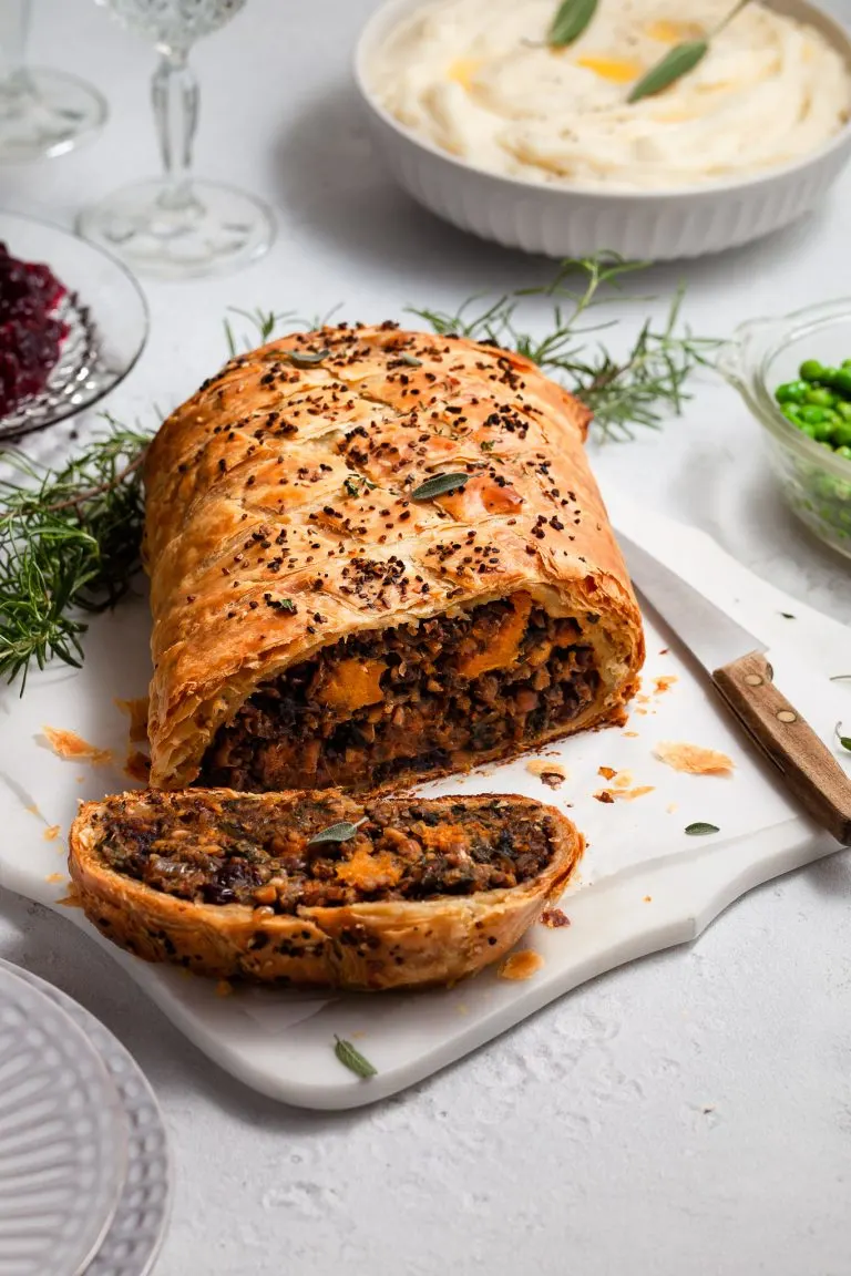 A vegan Wellington on a white marble platter sitting on a tabelscape with a bowl of whipped potatoes, green peas, cranberry sauce and wine glasses.