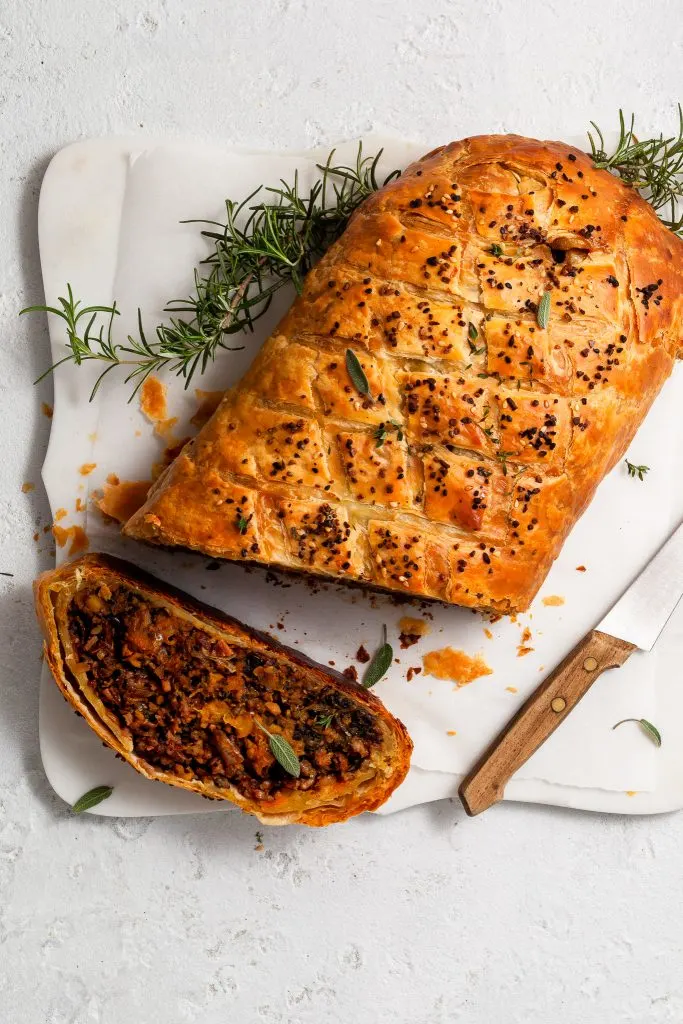Flatlay of a baked Vegan Wellington on a marble cutting board and piece sliced